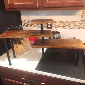 May include: A three-tiered serving stand with a dark wood finish and black metal supports. The stand has a rectangular shape and is designed for displaying food or decorative items. The stand is on a white countertop.