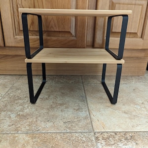 Könnte beinhalten: Ein zweistöckiges Regal mit einer hellen Holzoberfläche und schwarzen Metallstützen. Die rechteckigen Regale werden von schwarzen, eckigen Metallrahmen getragen. Das Regal ist für die Aufbewahrung oder Präsentation konzipiert.