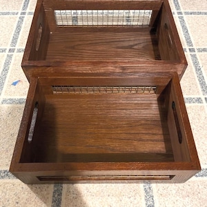 Könnte beinhalten: Zwei Holz-Aufbewahrungstabletts, die übereinander gestapelt sind. Die Tabletts sind in einem satten Braunton gehalten und haben einen Drahtgittereinsatz. Jedes Tablett hat ausgeschnittene Griffe an den Seiten.