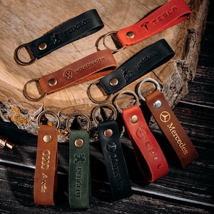 Op de afbeelding: Een verzameling leren sleutelhangers in verschillende kleuren, waaronder zwart, rood, bruin en groen. Elke sleutelhanger heeft een ander autologo, zoals Tesla, Peugeot en Mercedes. De sleutelhangers hebben een metalen ring en een drukknoopsluiting.