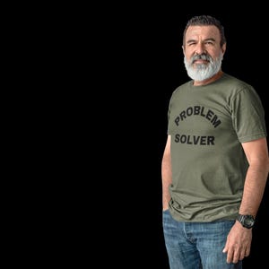 Könnte beinhalten: Ein olivgrünes T-Shirt mit dem schwarzen Aufdruck „PROBLEM SOLVER“. Die Person trägt eine blaue Jeans und eine schwarze Uhr. Der Hintergrund ist einfarbig schwarz.