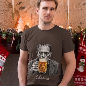 May include: A brown short-sleeve t-shirt featuring a graphic print of Frankenstein's monster holding a glass mug of beer with foam. Below the illustration, the word Cheers is printed in a white cursive script font on the front of the shirt.
