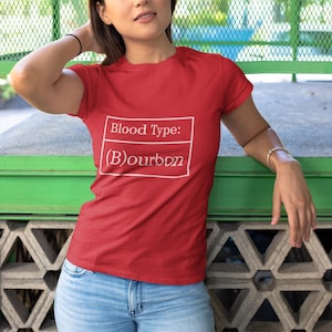 May include: Red t-shirt with the text "Blood Type: (B)ourbon" in a white box. The shirt is short-sleeved and is being worn with blue jeans. The background includes a green metal fence and a concrete wall.