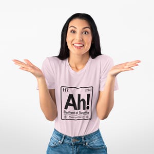 Op de afbeelding: Een lichtroze T-shirt met korte mouwen en een zwarte opdruk in de stijl van een element uit het periodiek systeem. De afbeelding toont het getal 117, de tekst Ah!, de zin Element of Surprise en de tekst Always unexpected.