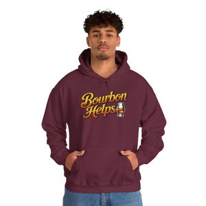 Può includere: Una felpa con cappuccio color bordeaux con la scritta Bourbon Helps in un carattere sfumato giallo e arancione, accompagnata da un'immagine di un bicchiere pieno di liquido ambrato. La felpa include una tasca a marsupio e lacci abbinati.
