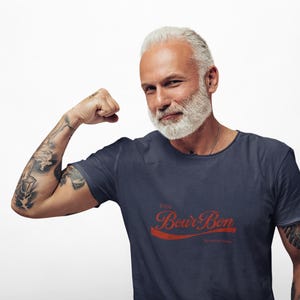 Könnte beinhalten: Marineblaues T-Shirt mit dem Text "Enjoy Bourbon" in roter Schrift. Das T-Shirt wird von einer Person mit grauem Bart und Tätowierungen am Arm getragen.