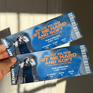 Könnte beinhalten: Zwei Konzertkarten für Billie Eilishs "Hit Me Hard and Soft" Tour, mit blau-orangem Design. Die Karten zeigen Veranstaltungsort, Datum, Uhrzeit, Bildschirm, Reihe und Sitzplatz. Die Tickets werden von einer Person gehalten.