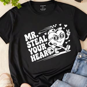 May include: Black t-shirt with a white graphic of a heart wearing sunglasses and holding a small bag, with the text "MR. STEAL YOUR HEART". The shirt has a crew neck and short sleeves.