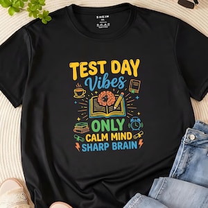 Puede incluir: Camiseta negra con un diseño colorido que dice "TEST DAY Vibes" con un libro abierto, un cerebro, un lápiz y otros iconos escolares. También incluye el texto "ONLY CALM MIND SHARP BRAIN".