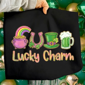 Puede incluir: Bolso tote negro con gráficos del Día de San Patricio. El diseño incluye un arcoíris sobre una olla de oro, una herradura con tréboles, un sombrero verde y una jarra de cerveza verde. Las palabras "Lucky Charm" están escritas debajo.