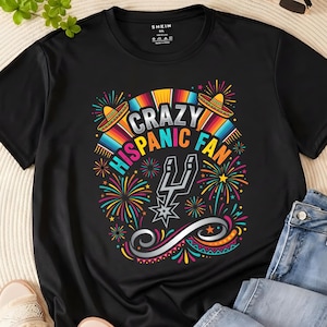 May include: Black t-shirt with a colorful graphic design. The design includes the text "CRAZY HISPANIC FAN" with sombreros, fireworks, and a silver spur logo. The shirt is a casual wear item.