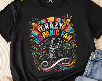 May include: Black t-shirt with a colorful graphic design. The design includes the text "CRAZY HISPANIC FAN" with sombreros, fireworks, and a silver spur logo. The shirt is a casual wear item.