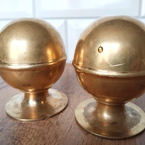 Puede incluir: Dos objetos decorativos antiguos de latón. Cada uno tiene una parte superior esférica y una base de pedestal. El latón tiene un tono dorado cálido, y los objetos parecen ser vintage. Se exhiben sobre una superficie de madera.