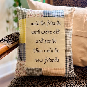 May include: A square patchwork throw pillow featuring a central cream-colored fabric panel with the embroidered black text: we'll be friends until we're old and senile then we'll be new friends. The border consists of various plaid and floral fabric squares.