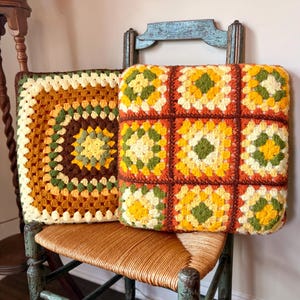 May include: Two square crochet pillows featuring granny square patterns in shades of yellow, orange, green, and cream. One pillow displays a single large central motif, while the other is composed of nine smaller joined granny squares with dark brown borders.