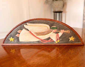Puede incluir: Una placa de pared de madera semicircular que presenta un ángel tallado en vuelo sobre un fondo gris oscuro. El ángel tiene alas de color crema, una túnica blanca con cintas rojas y toca una trompeta dorada. Dos estrellas amarillas flanquean al ángel.