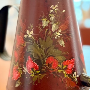 Könnte beinhalten: Eine maroonfarbene Metall-Teekanne mit Deckel, Griff und Ausguss. Die Teekanne ist mit handgemalten Erdbeeren, weißen Blumen und grünen Blättern verziert. Das Design befindet sich auf der Vorderseite.