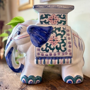 May include: A white ceramic elephant garden stool featuring hand-painted blue, green, and pink floral and scroll patterns. The elephant has a flat top surface, a trunk curled to the side, and a decorative saddle blanket with tassels along the bottom edge.