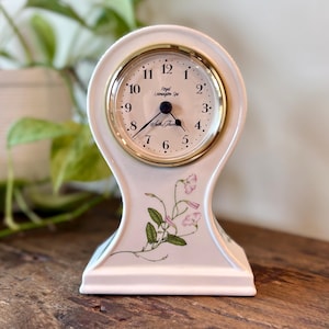 May include: A white ceramic mantel clock with a gold-toned circular bezel and black hands. The clock face features the text Royal Leamington Spa and Seth Thomas. A delicate botanical illustration of pink flowers and green leaves adorns the base.