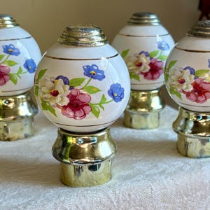 May include: Four decorative finials with a white, spherical design. Each finial features a gold base and top, with a floral pattern of red, blue, and white flowers. The finials are arranged on a white surface.