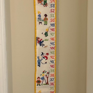 May include: A vintage embroidered height chart with a yellow border. The chart features embroidered figures and numbers from 27 to 60 inches. The figures depict children and various activities, adding a whimsical touch to the design.