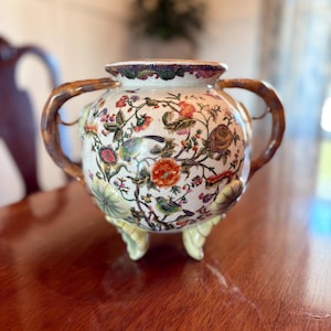 Könnte beinhalten: Eine dekorative Keramikvase mit cremefarbenem Sockel, verziert mit bunten Blumen- und Vogelmustern. Die Vase hat zwei braune Griffe und steht auf drei hellgrünen Füßen. Der Rand hat eine detaillierte Bordüre.