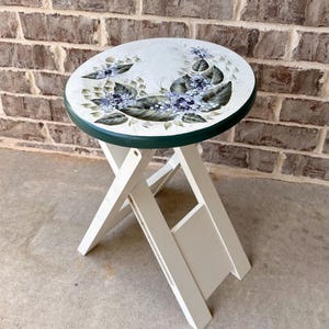 May include: A folding stool with a white frame and a round seat featuring a hand-painted design of blue hydrangea flowers and green leaves. The edge of the seat is painted a dark green, and the legs are connected by a white cross-brace structure.