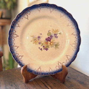 May include: A vintage porcelain plate with a scalloped edge and a blue border. The plate features a floral design in the center with yellow, purple, and green flowers. The plate is displayed on a wooden stand.