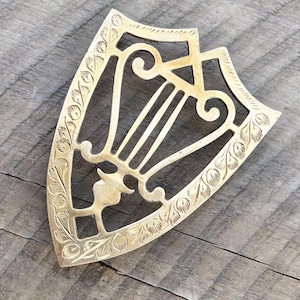 May include: A gold-colored, shield-shaped trivet with an ornate design. The trivet features a central harp-like pattern and a decorative border of leaves and swirls. The metal trivet is designed to protect surfaces from hot cookware.