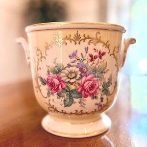 May include: Cream-colored ceramic planter with a floral design featuring pink roses, purple flowers, and green leaves. The planter has gold trim and handles, and sits on a pedestal base. A decorative piece for home decor.
