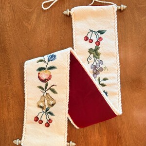May include: Two embroidered wall hangings with fruit designs. One features a peach, pears, and cherries, while the other displays grapes and cherries. The hangings have a cream-colored background, red velvet lining, and rope trim.