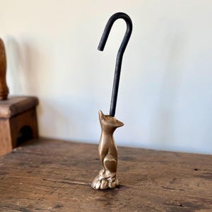 May include: A bronze-colored cat-shaped hook with a black, curved metal hook extending upwards. The cat figurine has a detailed, textured surface and is positioned on a wooden surface. The hook is likely for hanging items.