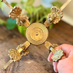 Peut inclure: Un support en métal doré doté d'une plaque centrale circulaire à anneaux concentriques et de quatre bras extensibles. Chaque bras se termine par un pied décoratif en forme d'éventail avec une petite tige métallique, au fini patiné.