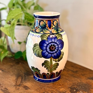 May include: A ceramic vase with a cream-colored base featuring hand-painted floral motifs in deep blue, green, and orange. The design includes a large central blue flower with green leaves and decorative border patterns at the neck and base.