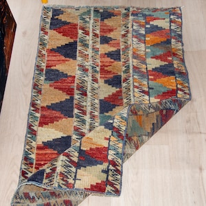 May include: A rectangular woven rug featuring a repeating geometric pattern of triangles in shades of navy blue, deep red, tan, and muted orange, with vertical striped borders and one corner folded over to reveal the underside texture.