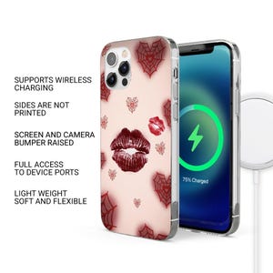 May include: A phone case with a light pink background featuring red spiderwebs, hearts, and lipstick kiss marks. The phone screen shows a charging symbol. The case supports wireless charging and has full access to device ports.