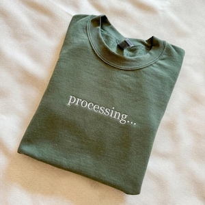 May include: An olive green sweatshirt with the word "processing..." embroidered in white. The crew neck sweatshirt is folded on a cream-coloured surface.