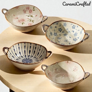 Può includere: Quattro ciotole in ceramica con manici, ognuna con motivi floreali o geometrici unici nei toni del blu, rosa e bianco. Le ciotole hanno un aspetto rustico e artigianale, con un bordo e manici marroni. Il testo "CeramiCrafted" è visibile in alto a destra.