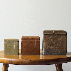 Puede incluir: Tres cajas cuadradas de cerámica con tapas en tonos marrones y verdes, sobre una mesa de madera. Las cajas tienen textura, la más pequeña con un diseño de hoja. Las cajas son de diferentes tamaños.