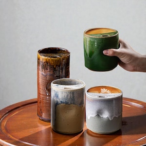 Könnte beinhalten: Eine Sammlung von Keramik-Kaffeetassen in verschiedenen Farben und Ausführungen. Die Tassen umfassen eine hohe, braune, zylindrische Tasse mit getropfter Glasur und mehrere kürzere, runde Tassen in Grün, Blau und Grau. Eine Tasse wird gehalten.
