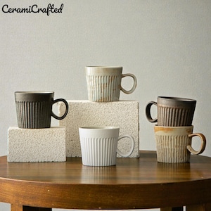 Könnte beinhalten: Eine Sammlung von Keramikbechern in verschiedenen Farben, darunter Weiß, Braun und Beige, mit vertikalen Rippendetails. Die Becher sind auf einem Holztisch und strukturierten Blöcken angeordnet. Der Markenname "CeramiCrafted" ist oben links sichtbar.