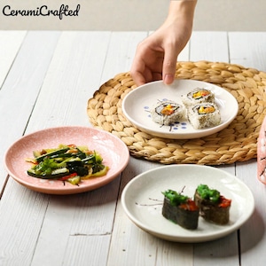 May include: A selection of ceramic plates, each with sushi rolls and vegetables. The plates are in pink and white, with a woven placemat in the background. The image is taken from a top-down perspective, with a hand reaching for a plate.