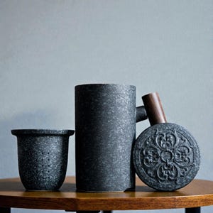 May include: A set of three dark gray ceramic tea accessories. The set includes a small tea strainer, a tall mug with a handle, and a round tea caddy with a wooden handle. The items are arranged on a wooden table.