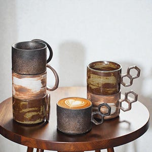 May include: Several ceramic mugs in brown and black tones, some stacked. One mug has a distinctive honeycomb-shaped handle. A single mug contains a latte with a heart design. The mugs are displayed on a wooden table.