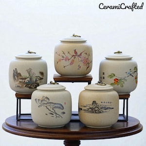 Puede incluir: Cinco tarros de cerámica beige con tapas, cada uno adornado con un diseño único de inspiración asiática pintado a mano. Los tarros presentan ilustraciones de paisajes, pájaros y flores. El texto "CeramiCrafted" es visible en la esquina superior derecha.