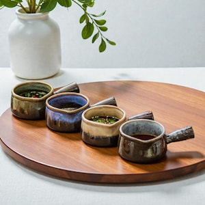 Puede incluir: Cuatro cuencos pequeños de cerámica para salsa con asas, cada uno lleno de una salsa diferente. Los cuencos son de tonos marrón, azul y beige, y están sobre una bandeja redonda de madera. Un jarrón blanco con hojas verdes está en el fondo.