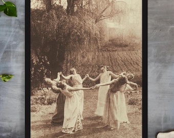 May include: A sepia-toned framed print depicts five people in flowing dresses holding hands in a circle, dancing in a field. The image has a vintage aesthetic, with a tree and a field in the background. The print is displayed on a wooden surface.