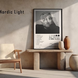 May include: A framed black and white poster featuring Mac Miller's album "Circles" displayed on a wooden table with decorative vases. The poster includes a portrait of the artist and a tracklist. A wooden chair sits to the left.