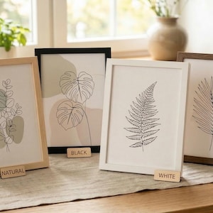 May include: Four framed botanical art prints with minimalist line drawings of leaves and plants. Frames are in natural, black, white, and walnut finishes. Each frame has a small wooden label with the frame color. The artwork is displayed on a wooden table.