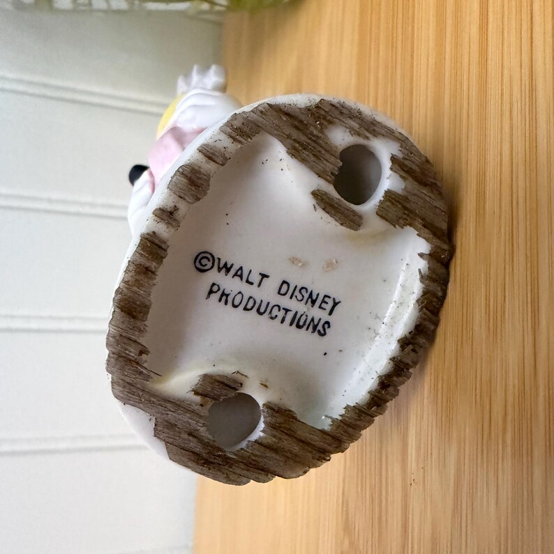 May include: A vintage ceramic figurine base with the text "Walt Disney Productions" printed in black. The base is white with a textured, brown rim and two holes. The figurine is partially visible, featuring pink and yellow accents.
